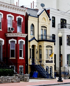 A row of colorful urban townhouses with intricate architectural details. The central building features a light yellow facade with dark blue accents and a flag hanging near the entrance. A staircase leads to the front door, and there is a lamppost nearby. Next to it, there are other buildings with red and white exteriors with similar decorative elements.