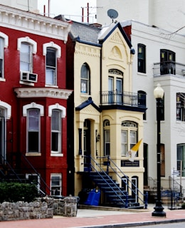 A row of colorful urban townhouses with intricate architectural details. The central building features a light yellow facade with dark blue accents and a flag hanging near the entrance. A staircase leads to the front door, and there is a lamppost nearby. Next to it, there are other buildings with red and white exteriors with similar decorative elements.