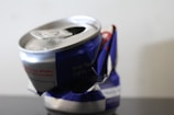 A crumpled aluminum can with a blue and silver design, featuring text and a partially visible logo. The top is open, with the tab still attached, and the can appears empty.
