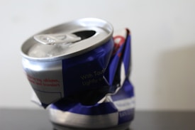 A crumpled aluminum can with a blue and silver design, featuring text and a partially visible logo. The top is open, with the tab still attached, and the can appears empty.