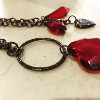 A close-up view of a piece of jewelry with dark metal chains and vibrant red gems. The gems have a polished, faceted surface, giving them a bright and lustrous appearance. The metal chain features a circular link and heart-shaped detail.