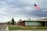 An American flag waves on a flagpole in front of a building with large windows and a brick exterior. The building is situated on a street corner, with a sign indicating 'Freedom Blvd'. The sky is overcast with gray clouds, suggesting a cloudy day. Trees and some distant hills can be seen in the background.