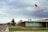 An American flag waves on a flagpole in front of a building with large windows and a brick exterior. The building is situated on a street corner, with a sign indicating 'Freedom Blvd'. The sky is overcast with gray clouds, suggesting a cloudy day. Trees and some distant hills can be seen in the background.