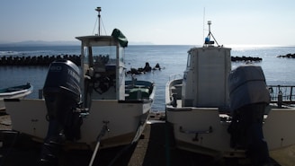 Two motorized boats are parked at a dock by the sea. The boats display visible engines and are positioned side by side. The calm sea stretches out in the background, with a line of breakwater visible and a faint outline of distant mountains.