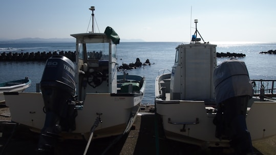 Two motorized boats are parked at a dock by the sea. The boats display visible engines and are positioned side by side. The calm sea stretches out in the background, with a line of breakwater visible and a faint outline of distant mountains.
