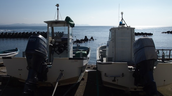 Two motorized boats are parked at a dock by the sea. The boats display visible engines and are positioned side by side. The calm sea stretches out in the background, with a line of breakwater visible and a faint outline of distant mountains.