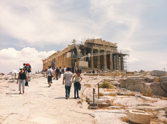 A group of people walking towards an ancient temple structure partially covered in scaffolding. The temple is situated on rocky terrain under a bright, partly cloudy sky. Some of the people are taking photos and wearing casual, summer attire.