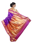 A woman is elegantly draped in a traditional saree with vibrant colors and intricate patterns. The saree features a rich combination of purple and gold with decorative motifs, including peacocks and floral designs. Her hair is styled in an elegant updo, and she accessorizes with earrings.