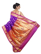 A woman is elegantly draped in a traditional saree with vibrant colors and intricate patterns. The saree features a rich combination of purple and gold with decorative motifs, including peacocks and floral designs. Her hair is styled in an elegant updo, and she accessorizes with earrings.