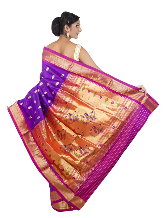 A woman is elegantly draped in a traditional saree with vibrant colors and intricate patterns. The saree features a rich combination of purple and gold with decorative motifs, including peacocks and floral designs. Her hair is styled in an elegant updo, and she accessorizes with earrings.