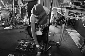 A musician kneels on stage, adjusting effects pedals with a guitar slung around their neck. The setting is filled with musical equipment including drums, an amplifier, and sound cables. The image is in black and white, contributing to a vintage or classic concert feel.