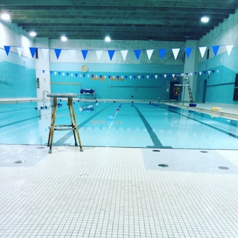 An indoor swimming pool with clear, calm water. White and blue triangular flags hang overhead across the pool. A solitary wooden stool is placed at the edge of the pool area. The walls and ceiling are painted in various shades of blue. On the far wall, there's a clock and some decorations. Several pool accessories, such as floats, are scattered around.