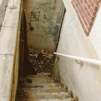 A narrow concrete stairway leading down to a basement area, flanked by brick and concrete walls. Graffiti in various colors is visible on the walls. The steps are worn and covered with dirt and debris. At the bottom, there are scattered items including pieces of wood, a broken pallet, and some trash.