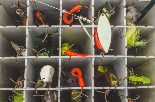 A tackle box is organized with various fishing lures and hooks in separate compartments. The lures include bright red and green soft plastics, along with shiny spoon lures and other fishing accessories. The compartments are neatly divided, with different types of gear in each section.