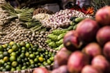 A vibrant display of various fresh vegetables and fruits, including green limes, lemongrass, cucumbers, bitter gourds, onions, and red shallots. The produce is arranged in woven baskets, creating a colorful and abundant market scene.