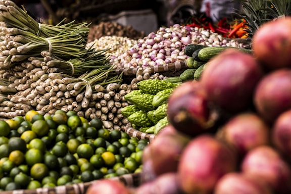 A vibrant display of various fresh vegetables and fruits, including green limes, lemongrass, cucumbers, bitter gourds, onions, and red shallots. The produce is arranged in woven baskets, creating a colorful and abundant market scene.