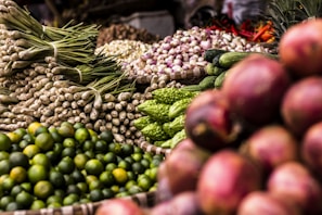 A vibrant display of various fresh vegetables and fruits, including green limes, lemongrass, cucumbers, bitter gourds, onions, and red shallots. The produce is arranged in woven baskets, creating a colorful and abundant market scene.
