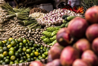 A vibrant display of various fresh vegetables and fruits, including green limes, lemongrass, cucumbers, bitter gourds, onions, and red shallots. The produce is arranged in woven baskets, creating a colorful and abundant market scene.