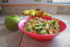 A vibrant salad composed of chopped cucumbers, tomatoes, and leafy greens is presented in a bright red bowl placed on a marble countertop. Nearby, a green apple and a bunch of bananas add to the fresh and healthy theme of the arrangement.