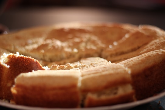 A freshly baked, round cake with a golden-brown crust is placed on a plate. The cake is partially sliced, revealing a soft and fluffy texture inside. The edges are slightly darker, indicating a crisp finish, and the lighting creates a warm and inviting atmosphere.