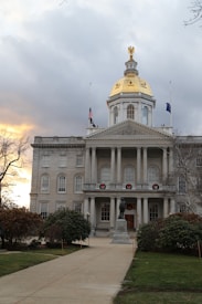 A large government building with a golden dome at the center of its roof. The building has multiple rectangular windows and is adorned with columns at the entrance. Two flags are flying atop the structure. A statue stands before the entrance on a stone pathway flanked by trees and shrubs. The sky is partly cloudy with a hint of sunset light.