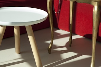 A small, round, white table with wooden legs stands next to a taller, golden table with curved legs. The furniture is set against the backdrop of a red sofa.