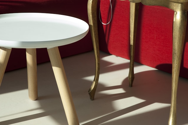 A small, round, white table with wooden legs stands next to a taller, golden table with curved legs. The furniture is set against the backdrop of a red sofa.