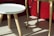 A small, round, white table with wooden legs stands next to a taller, golden table with curved legs. The furniture is set against the backdrop of a red sofa.