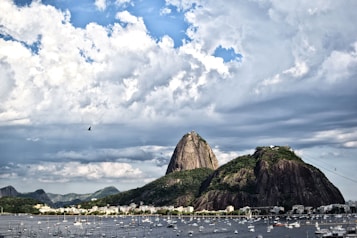 A stunning view of the famous Sugarloaf Mountain with its unique shape, rising prominently above the landscape. The foreground features a calm sea with numerous small boats moored, and a cityscape extending along the shoreline. The sky is filled with dramatic clouds, creating a picturesque setting.