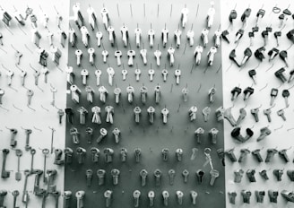 An assortment of various keys organized in rows and columns, hanging on hooks on a display board. The keys are metallic and vary in shapes and sizes, indicating they may serve different locks or purposes. The background is a combination of light and dark tones, contributing to a minimalistic and organized aesthetic.