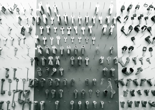 An assortment of various keys organized in rows and columns, hanging on hooks on a display board. The keys are metallic and vary in shapes and sizes, indicating they may serve different locks or purposes. The background is a combination of light and dark tones, contributing to a minimalistic and organized aesthetic.