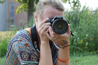 A person with blonde hair is holding a Canon camera up to their face, taking a photograph. They have multiple rings on their fingers and a tattoo on their wrist. The individual is wearing a colorful, patterned shirt and a black camera strap around their neck. In the background, there is greenery including grass and trees, suggesting an outdoor setting.