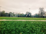 A group of people are playing soccer on a green artificial turf field. The scene is set outdoors, with several players in motion and a soccer goal in the background. The sky is overcast, and there are trees and a fence surrounding the field.