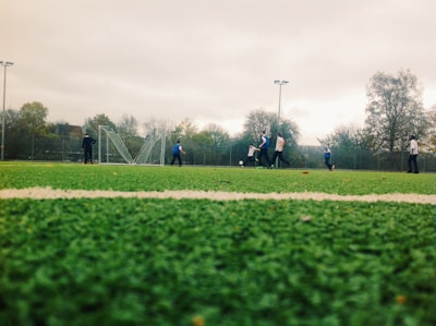 A group of people are playing soccer on a green artificial turf field. The scene is set outdoors, with several players in motion and a soccer goal in the background. The sky is overcast, and there are trees and a fence surrounding the field.