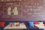 A cozy cafe interior features a mural on the wall with whimsical handwritten text in a foreign language and illustrations of a couple having coffee. The seating area includes a plush couch with colorful pillows in red, blue, and yellow. On the wooden tables, there are glasses, a red ashtray, and a few ceramic mugs.
