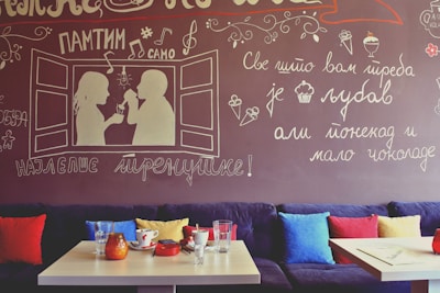 A cozy cafe interior features a mural on the wall with whimsical handwritten text in a foreign language and illustrations of a couple having coffee. The seating area includes a plush couch with colorful pillows in red, blue, and yellow. On the wooden tables, there are glasses, a red ashtray, and a few ceramic mugs.