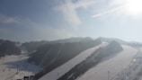 A snowy landscape with multiple ski slopes surrounded by forested hills. The slopes are lined with chairlifts and dotted with skiers. The horizon is hazy under a bright, possibly midday sun shining through a partly cloudy sky.