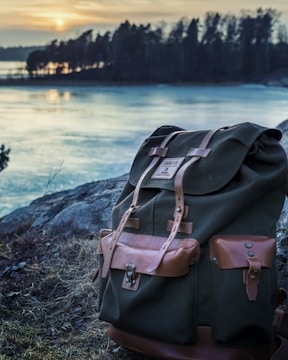 A rugged backpack with leather straps and pockets sits on a rocky terrain near a calm lake with a forest in the background. The sun is setting on the horizon, casting a warm glow over the scene.