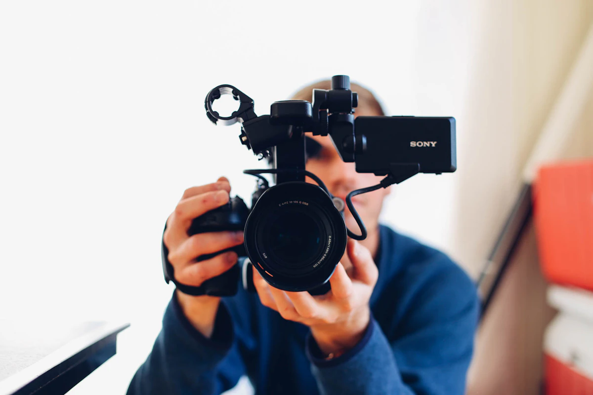 Un opérateur tient une caméra vidéo professionnelle à hauteur de visage, les deux mains posées sur l'objectif, dans une pièce claire