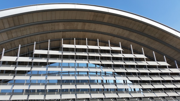 An architectural structure featuring a series of horizontal louvered panels arranged in an arc. The design allows for both functionality and aesthetics, with metallic elements and large glass windows reflecting the sky. The structure appears modern and utilitarian.