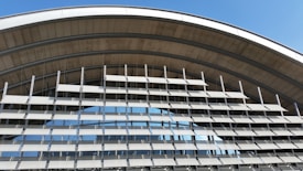 An architectural structure featuring a series of horizontal louvered panels arranged in an arc. The design allows for both functionality and aesthetics, with metallic elements and large glass windows reflecting the sky. The structure appears modern and utilitarian.