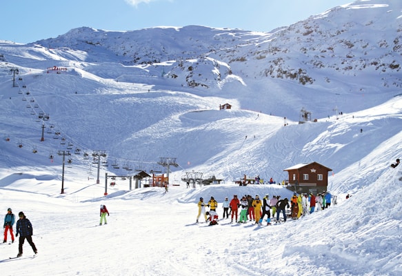 Sunny day at a ski resort with snow-covered mountains in the background. People dressed in colorful ski outfits are either skiing, walking, or standing in groups near a small wooden building. A series of ski lifts run up the slopes, carrying skiers to higher elevations.