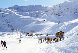 Sunny day at a ski resort with snow-covered mountains in the background. People dressed in colorful ski outfits are either skiing, walking, or standing in groups near a small wooden building. A series of ski lifts run up the slopes, carrying skiers to higher elevations.