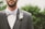 A man dressed in a formal suit featuring a gray jacket and white shirt. He wears a striped bow tie and a boutonniere with feathers and berries. The background is blurred greenery.