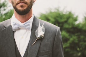 A man dressed in a formal suit featuring a gray jacket and white shirt. He wears a striped bow tie and a boutonniere with feathers and berries. The background is blurred greenery.