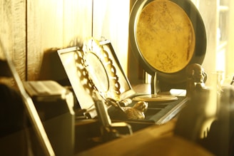 A collection of antique or vintage items arranged on a wooden shelf. Prominent objects include a decorative gold plate with intricate floral engravings, an ornate picture frame, and a small statuette of a person. The warm, golden hue lighting suggests a cozy, nostalgic atmosphere.