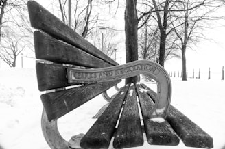 A wooden bench partially covered in snow is situated in a wintry park setting. The words 'Parks and Recreation' are engraved on the metal side arm of the bench. Bare trees are visible in the background, with a snow-covered ground and a few lamp posts lining the area.