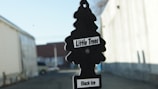A black, tree-shaped car air freshener labeled 'Little Trees' and 'Black Ice' is hanging in front of a blurred outdoor background. The setting appears to be an alley or street with buildings on either side.