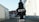 A black, tree-shaped car air freshener labeled 'Little Trees' and 'Black Ice' is hanging in front of a blurred outdoor background. The setting appears to be an alley or street with buildings on either side.