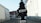 A black, tree-shaped car air freshener labeled 'Little Trees' and 'Black Ice' is hanging in front of a blurred outdoor background. The setting appears to be an alley or street with buildings on either side.